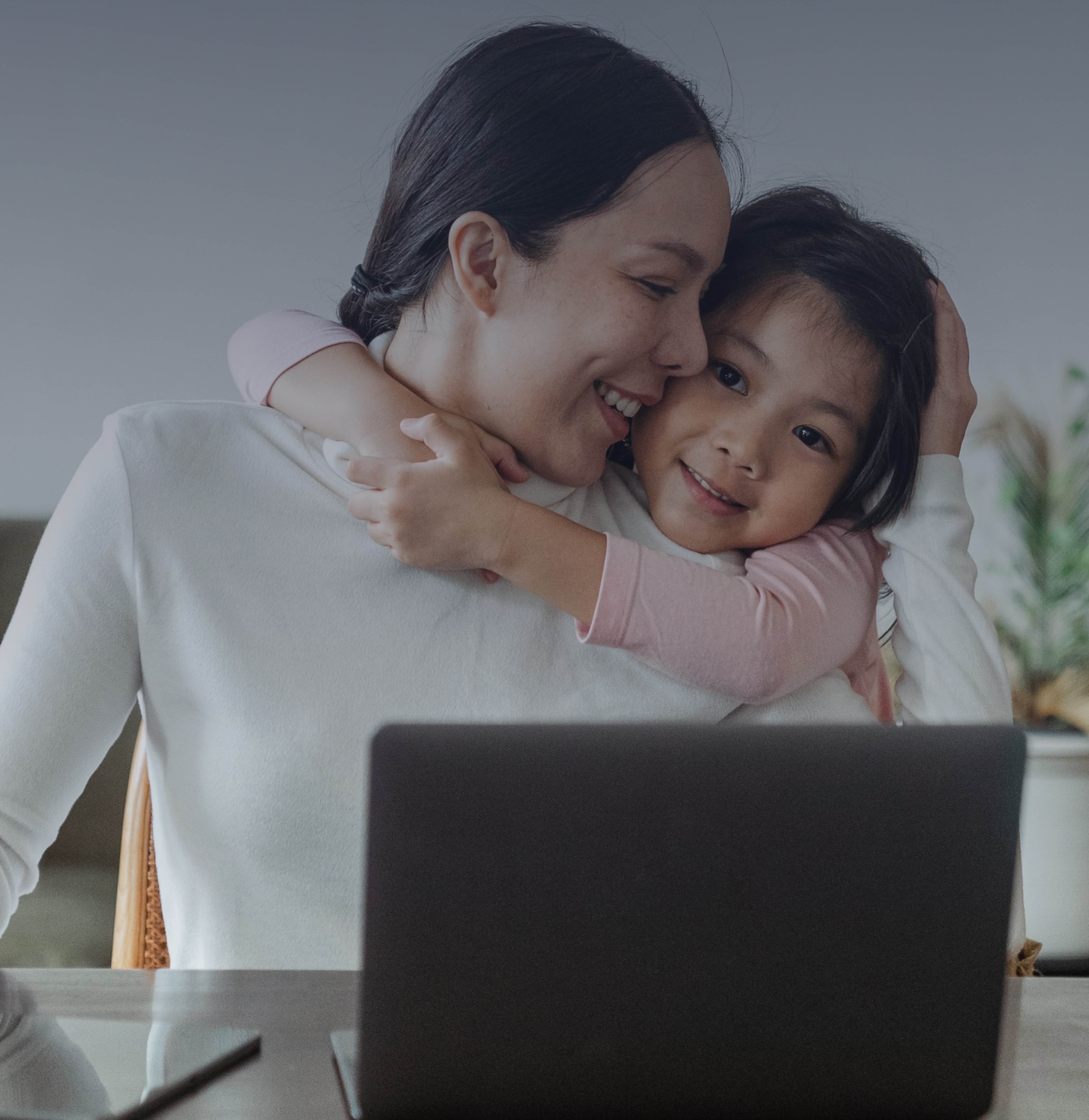 A young girl hugging her mother while she works at a laptop