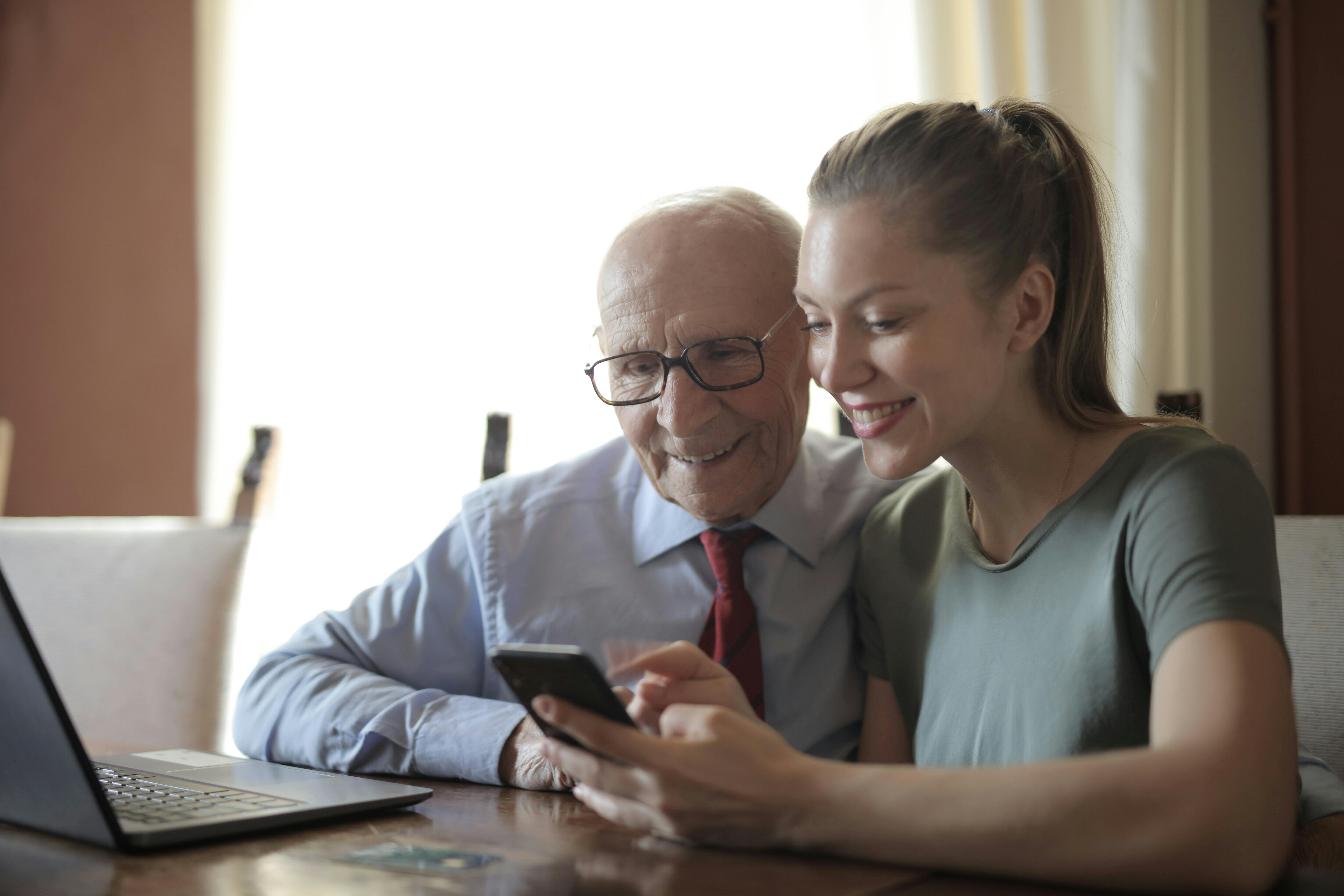 Older man and younger woman smiling and sitting at a table, looking at a mobile phone with a laptop open on the table
