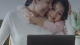 A young girl hugging her mother while she works at a laptop